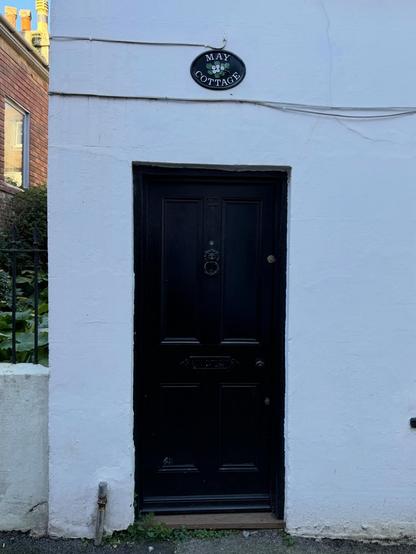 A black wooden front door set into the corner of a white building, a plaque above the door reads "May cottage", and also in black in the door is the number 11.