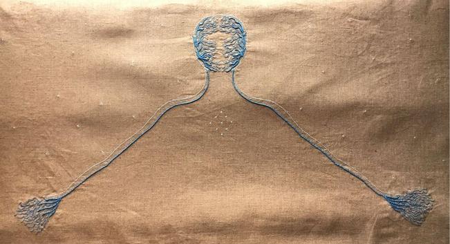 An embroidery on ochre linen, there is the outline of a head and outstretched arms. Tiny blue filigree decorates the face and hands and there are light coloured stars on the chest area and around the neck.&nbsp;
