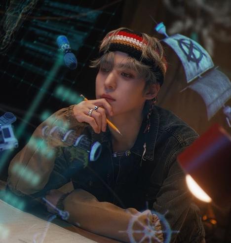 Ateez Yunho dans le MV de Crazy Form. 
A tout l’air assis, appuyé contre une table, ses deux bras posés sur la surface et appuie sa tête sur sa main droite qui tient un crayon jaune en ayant l’air pensif et concentré. 
A les cheveux gris clairs qui lui tombent sur les yeux et porte un bandeau sur le front. 

On peut voir en transparence devant et autour de lui une sorte d’interface numérique inspiration de l’imagerie du code et de la programmation à la fois rétro et futuriste.
Or celle-ci fait un poil plus de sens puisqu’elle présente des images de certains éléments importants du lore de Ateez : un bateau à voile, un gouvernail…
Ces éléments semblent flotter tout autour de lui.