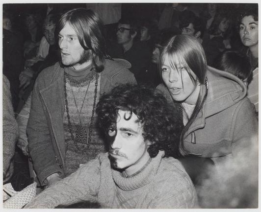 The black and white photograph features a group of people at what appears to be an indoor gathering or event. In the foreground, there is a man with curly hair wearing a turtleneck sweater looking directly towards the camera; his expression is serious and contemplative. Beside him stands a woman in profile view who seems engaged in conversation with another individual not fully visible in this frame. She has long straight hair parted down the middle, wears glasses perched on her nose bridge, and appears to be wearing a coat over a turtleneck sweater or dress.

In the background, several other individuals are partially visible; some appear attentive while others seem distracted by something outside of the camera's view. The crowd is dressed in winter clothing such as coats, scarves, and knitwear, suggesting that this gathering took place during colder months. There's a sense of movement captured with people looking around or engaging in various activities.

The image has an overall candid quality to it, capturing what seems like spontaneous interactions among the attendees at a public event without any apparent staging or formal composition. The absence of color and focus on attire suggest that this could be from a historical context, perhaps connected to social movements as indicated by the reference "Anti Apartheid protest" in the alt text provided.

The image's vintage  [...]
