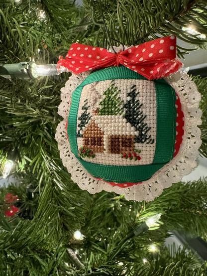 A tree ornament that is round, but flat and slightly stuffed. It has white lace around the edge, then red with white polka dot fabric followed by green grosgrain ribbon. In the center is a square that is needlepoint. It has a white background, a brown cabin with white roof, smoke coming from the chimney and pine trees in the background. On top of the ornament is a red bow that has white polka dots. It’s hanging on a tree.