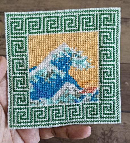a rendition of the great wave off kanagawa,  with a yellow sky and blue,green, white threads to design the wave. 

there is a green border designed all around.