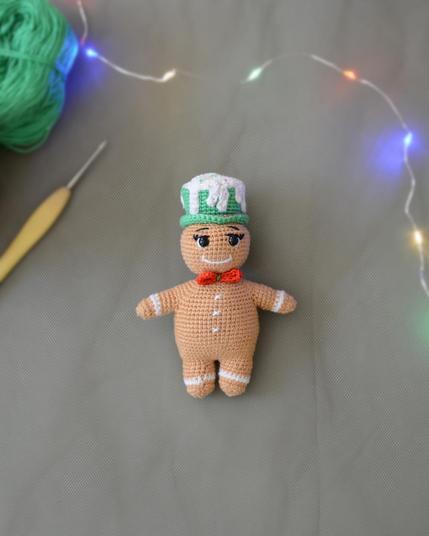 Petit bonhomme en pain d'épices au crochet posé au centre d'un fond uni. Il porte un nœud papillon rouge, un chapeau vert décoré d'un "glaçage" blanc et a des détails blancs sur les bras, les jambes et le torse. En haut de l'image, une guirlande lumineuse colorée floue apporte une ambiance douce et festive. En haut à gauche, on voit une pelote de fil vert et un crochet.