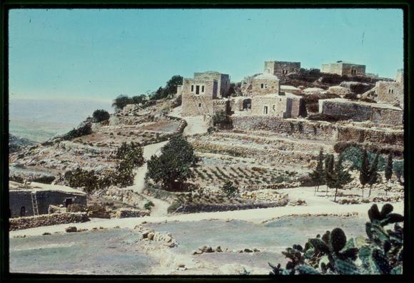This image depicts an ancient village situated on a hillside. The structures appear to be made of stone, with multiple levels ascending the slope, suggesting a defensive or strategic position. There are terraced fields carved into the terrain, indicative of agricultural practices and land management suited for arid conditions.
The landscape is dry with sparse vegetation, including some trees and what seems like cacti in the foreground on the right side. The sky above appears clear, hinting at a sunny climate. A sense of history and antiquity pervades the scene as there are no modern elements visible.
Given the photograph's attribution to "Matson Photo Service" and context provided by Jaffa to Jerusalem with reference between 1950 and 1977, it is likely that this image captures a historical or archaeological site in Israel. The specific mention of Upper Beth Horon village corresponds with biblical references from Joshua chapter sixteen verse five (Joshua 16:5).