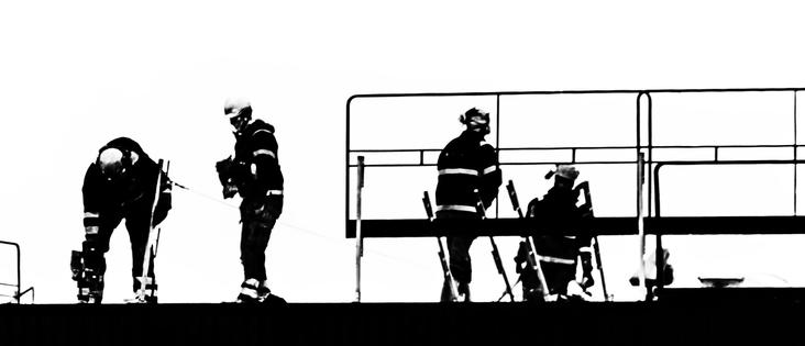 Four construction workers wearing helmets and safety gear working on a raised platform with tools and railings, silhouetted against a bright background.