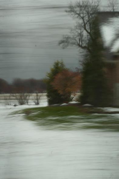 Color photo taken out the side window of a car while travleing about 35 mph, at maybe 1/10th sec shutter speed. The edge of a home pokes into the right side of the image. Next to it we see a well-captured evergreen tree and the ghostly apparition of some other tree that looks golden and floating. Bottom of scene is snow, streaked by motion. Top of the scene is similar--a dark grey overcast sky streaked with foreground trees that are merely streaky blurs passing by.