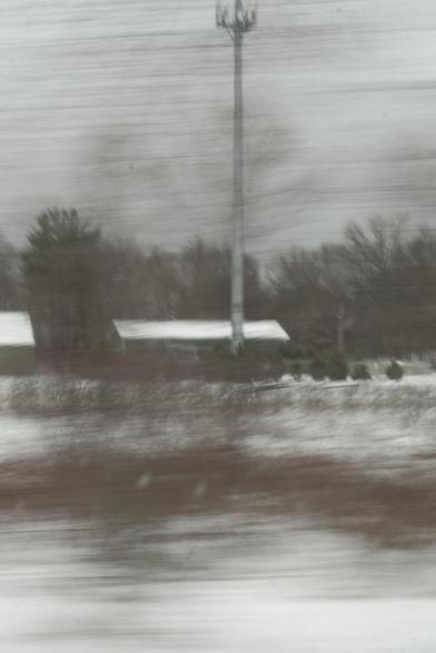 Blurry/streaked color photo of rural landscape. a snowy field in the foreground, two single-story structures in the midground, a cell phone tower rising above them, and a line of trees at the horizon. in the foreground, trees next to the road are rendered practically invisible by the car speed vs shutter speed, but the trees leave horizontal streaks that look like brush strokes across the image.