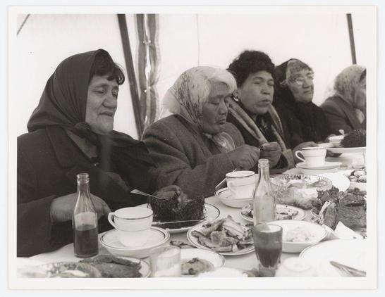 The photograph captures a group of individuals seated around an outdoor dining table, engaged in the act of eating. The scene appears to be set during daytime under natural light conditions. On the table are various dishes and food items such as meat, bread, salads, and what looks like cake or pastry slices on individual plates. Beverages include at least two bottles with dark contents that could possibly be soda or juice. Each person has a cup in front of them, some holding utensils like forks or spoons suggesting they are consuming the meal.

The individuals exhibit diverse expressions; one appears to be enjoying their food while another looks into space, perhaps lost in thought. The attire suggests it might not be warm weather as coats and scarves can be seen worn by several people at the table. There is a sense of casualness yet attentiveness towards each other's presence during this communal meal.

The black-and-white nature of the photo adds to its timeless feel, while the setting implies an outdoor or semi-outdoor environment due to what appears to be white fabric in the background that could suggest some form of sheltered area. The photograph carries a candid and unposed quality capturing a slice-of-life moment among these individuals.