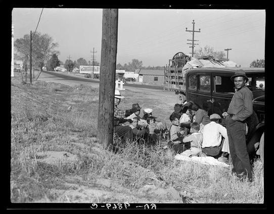 The black and white photograph depicts a group of individuals gathered near an older model car with luggage stacked on top. The setting appears to be rural, as evidenced by open fields, scattered trees, and utility poles in the background. A telephone pole is prominently positioned in the foreground.

Several people are seated or standing around the vehicle; some appear contemplative while others engage in conversation. Notably, there's a mix of adults and children within this group. The individuals exhibit diverse clothing styles typical for that era, with hats being common headwear among both men and women.

The overall atmosphere suggests a moment captured during travel or perhaps waiting to commence their journey. No direct interaction between the subjects is observable; they seem absorbed in individual thoughts or activities rather than engaging directly within this frame of time.