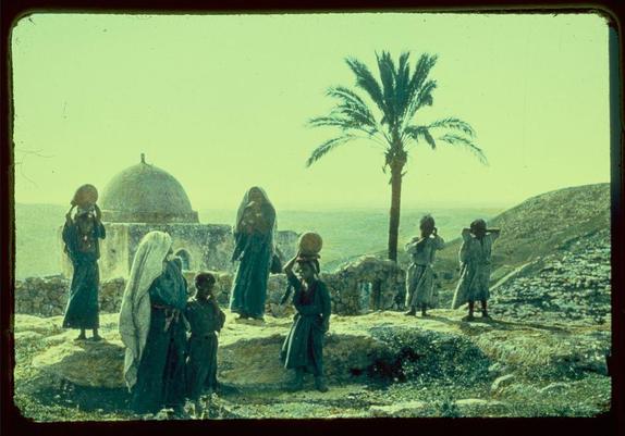 The image depicts a scene with several people standing outdoors, dressed in traditional Middle Eastern clothing. The setting appears to be an arid landscape, possibly on the edge of a historical site or ruins, given the presence of ancient architecture and a palm tree.

There are seven individuals visible: four women wearing long robes covered by head coverings (hijabs), two young boys standing nearby, and another woman with her back turned. The group is spread out across what looks like rocky terrain, possibly on elevated ground overlooking an expanse that could be the Mediterranean Sea or a similar body of water.

In the background stands a large dome structure, which might suggest a religious site such as a mosque. A palm tree sways in the wind behind this building, adding to the sense of arid climate and historical context. The overall colors are muted with earthy tones dominating the scene, complemented by a hazy sky.

The image is marked with greenish-yellow tint across it, which could be due to aging or processing effects on the photograph itself. At the bottom right corner, there's text that reads "Jaffa to Jerusalem Zorah Home of Samson Judges 13:2 between 1950 and 1977 Matson Photo Service" suggesting a possible connection to biblical themes from those regions during this time period.

The image is credited as belonging to the Eric & Edith Matson Photographs, sp [...]