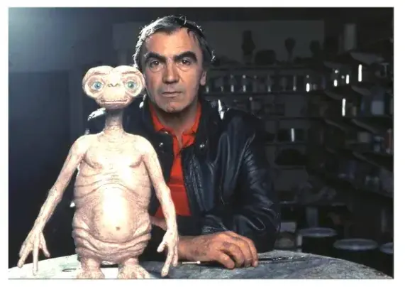A dark-haired white Italian man in a red shirt and a black leather jacket sits in a workshop with a slender paintbrush and a model of E.T.