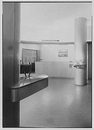 The image depicts an interior space, specifically a section of the First Federal Savings and Loan Association located in the RCA Building at Rockefeller Center in New York City. The photograph is taken on December 12th, 1944 and shows several modern design elements typical for that time period.
In this black-and-white photo, we can observe an expansive room with a polished concrete floor reflecting some light sources scattered around. A prominent feature of the space is a sleek, cantilevered reception desk on one side, likely intended to be unobtrusive and serve as an efficient communication point for visitors or personnel.
The area in front of the counter seems clean and minimalistic, with no clutter visible, suggesting that it might have been taken during off-hours or when not many people were present. On the right-hand side, a cylindrical structure, possibly part of a building's architectural design, stands tall near the room's back wall.
There are also other furniture pieces like chairs which add to the functionality and style of this space. The absence of natural light indicates that it is either an interior hallway or not directly exposed to sunlight coming through windows; instead, artificial lighting sources illuminate different parts of the room as seen in reflections on the floor.