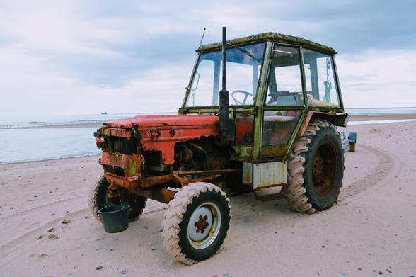 A photo of a rusted tractor on a beach. Its cab has a boxy look and the whole thing feels quite 1980s. Despite the shoddy bodywork, the tyres are covered in sand and the tracks behind it are brand new so this thing is still tractoring.