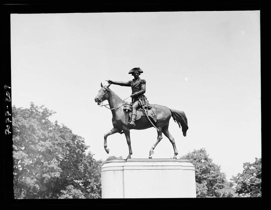 The image is a black and white photograph of an equestrian statue, depicting a person mounted on horseback. The rider appears to be dressed in historical military attire with a tricorn hat indicative of 18th-century fashion, possibly representing someone from the American Revolutionary War era or similar period. They are pointing towards something off-camera with their right hand while holding the reins in their left. The statue is positioned atop a tall pedestal set against an outdoor backdrop with trees and clear sky visible above it. There's no indication of movement as if caught mid-action; instead, there’s a sense of stillness conveyed by both the posture of rider and horse.