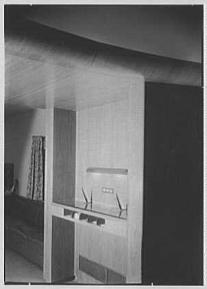 The image depicts a classic office setting from the mid-20th century, showcasing an elegant and functional workspace. The photograph captures two desks against opposite walls in what appears to be a modernist-style interior with clean lines and minimalistic design.

On one side of the room stands a sleek desk made of light-colored wood or laminate, equipped with several drawers for storage below and shelves above that hold books and office supplies such as telephones. The back wall features a large window draped with patterned curtains, allowing natural light to illuminate the space.

Adjacent to it is another similar yet unoccupied work area on the opposite side, emphasizing symmetry in the room's design. In between these two desks are several chairs arranged for meetings or collaborative discussions. A noticeable feature of this office environment includes its simplicity and focus on utility without excessive ornamentation—a hallmark of mid-century modern aesthetics.

Overall, the image reflects an era where practicality met style within professional environments while maintaining a sense of orderliness and sophistication through architectural choices and furniture selection.