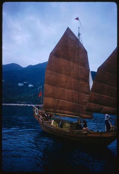 A traditional sailing vessel with large brown sails is seen on a body of water surrounded by hills and a partly cloudy sky. The ship has two visible masts, each adorned with several square sails made from an earthy-colored fabric. Flags are attached to the topmost mast, adding detail to this distinctive sailboat design. Onboard, there appear to be at least four people: one in white standing near the back of the boat under a shaded area, another wearing pink and black sitting on the deck closer to the stern, two more individuals partially visible; they are dressed casually with hats or head coverings.

The scene captures both human activity and natural beauty. The water is calm, reflecting light from various sources including what might be early evening sunlight that casts a warm hue over parts of the image. In the background, buildings on a hill suggest an urban environment not far away from this serene nautical setting. Overall, it's a picturesque moment captured in time by Toni Frissell during her assignment for Sports Illustrated.

Note: The date mentioned in your prompt is July 1959 which could be relevant to understanding the historical context of Hong Kong at that time as well as providing information about when this photograph was taken or intended.