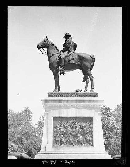 A black and white photograph of a bronze equestrian statue depicting a man in military uniform riding a horse. The rider is dressed in dark clothing, including a long coat and hat, with reins held loosely in his hands as he looks forward. Below the main figure, there's an intricate bas-relief depiction of soldiers marching in formation within a decorative frame on what appears to be a pedestal or plinth. The image has handwritten annotations at its bottom: "70 24-210" and is attributed to Arnold Genthe with dimensions noted as "5 x 6".