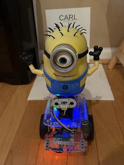 Robot Carl (with 8 inch tall, single eye Minion character Carl) atop the GoPiGo3 differential drive robot platform.  Carl's eye pupil is replaced by a PiCamera, and he has a microphone for speech recognition.  In the photo the floor glows from the LED on Carl's floor facing speaker which he uses for his text to speech communication.  He also has a forward facing time of flight range sensor that he uses to ensure he docks with millimeter accuracy.