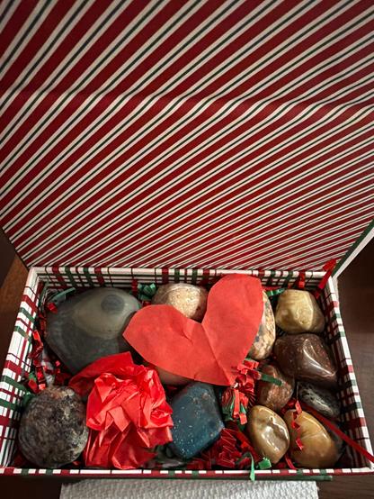 A holiday box filled with various stones that have been tumbled and polished with a red tissue heart.