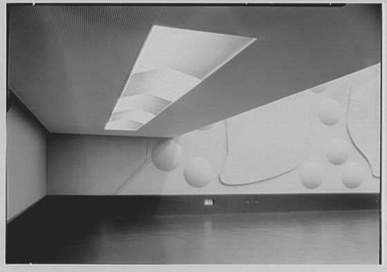 The image is a black and white photograph of an interior space with architectural features. The perspective shows the corner where two walls meet, forming a right angle. One wall has a series of relief sculptures or cutouts that create geometric shapes in three-dimensional form, including what appears to be circles and lines creating abstract patterns.

Above this feature wall is a large skylight or vented section on the ceiling, allowing light into the space. The surface below the windows appears smooth but without any discernible features like fixtures or objects placed on it.

The floor extends towards an empty open area with no visible furniture or people within that part of the room. It seems to be a large expanse for movement, possibly indicating this could be a gallery or exhibition hall given its minimalist and clean design aesthetic.

Overall, the photograph captures the starkness and simplicity of modernist interior architecture from the mid-20th century.