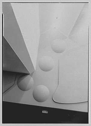 This black and white photograph depicts a modernist abstract sculpture composed of geometric shapes, including spheres and cones. The artwork is suspended in mid-air against an off-white background that appears to be part of its display setting.
The sculpture features several floating forms: two large spherical objects, one larger than the other; a smaller sphere above them; a cone-like shape pointing upward with open apex facing toward these spheres; another conical form lying on its side. The overall composition has an airy and minimalist quality suggesting it is part of a contemporary art exhibit.
The photograph captures this piece in great detail, highlighting the smooth textures and shadows cast by each object within the sculpture's shadowed recesses. This provides depth to their otherwise flat surfaces while creating contrast between light and dark areas throughout its structure.
In terms of context: National Broadcasting Co., Rockefeller Center refers specifically to NBC Studios at 30 Rock (Rockefeller Plaza) in New York City, which is part of the historic Rockefeller Center complex known for being home to major media companies. The date mentioned - October 23rd - might indicate when this photograph was taken or associated with an event related to it.
The reference "Gottscho-Schleisner" seems to point towards Gottschalks, Inc., a New York-based photography studio a [...]