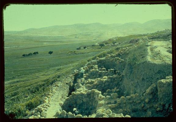 The image depicts a rural landscape with terraced agricultural fields extending into the distance. A prominent feature in the foreground is an archaeological excavation site, characterized by uneven ground and remnants of ancient structures made from stones or rocks piled up haphazardly. The terrain appears dry, indicative of arid conditions typical for the region.
Trees dot various points along the landscape, suggesting a semi-arid environment with pockets of vegetation. In the middle distance, more terraced fields can be seen alongside what seems to be small clusters of buildings and paths winding through them. Rolling hills form a backdrop under an overcast sky, creating soft contours across the horizon. This scene captures both historical exploration in terms of archaeology and agriculture amidst natural undulations.
This image has been provided as alt text for visually impaired users with additional context regarding its source from Jaffa to Jerusalem: Beth Shemesh section documenting excavations between 1950-77, by Matson Photo Service. The photo is a slide measuring approximately two inches square and presented in color.
