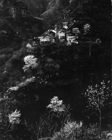 The image is a black and white photograph depicting a quaint village situated on the side of a hill. The structures are traditional, with multiple levels ascending up the incline, suggesting historical or rural significance. There's noticeable contrast between illuminated buildings and surrounding darkness, likely from artificial lighting within the homes as it appears to be nighttime or early evening given the low light conditions. Trees scattered throughout frame add depth and context, some of which appear in bloom while others are bare, hinting at a seasonal change possibly autumn or spring. The absence of visible people gives an impression of isolation or quietness. This photograph seems artistically captured rather than for documentary purposes, with its dramatic lighting emphasizing the textures and contours of both natural landscape and built environment.