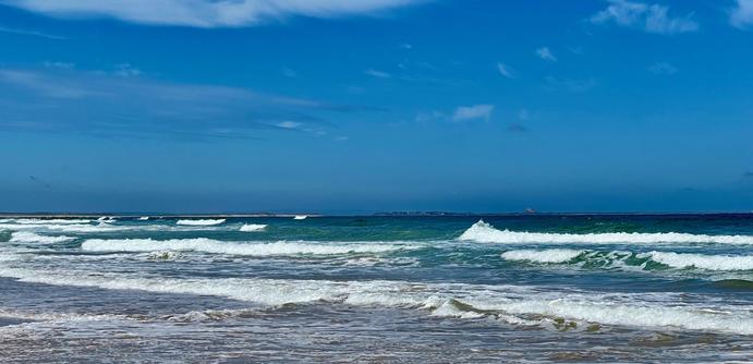 Waves roll gently towards a sandy shoreline under a bright blue sky with scattered, thin clouds. The sea shifts from deep blue to lighter turquoise near the shallows, where white foam forms as the waves break. The horizon stretches across the frame, with small, distant landforms barely visible against the water. The beach is wet near the edge, reflecting some of the sky’s colour, and the scene is calm with no people or activity present.