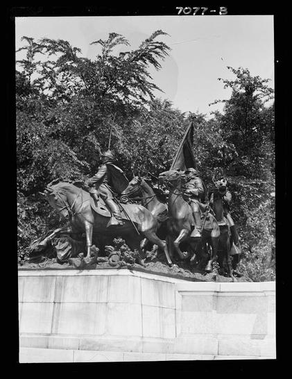 The image shows a black and white photograph of an equestrian statue depicting historical figures on horseback. The scene captures multiple horses and riders, with one figure prominently holding what appears to be a flag or banner aloft. There are several other individuals shown in various poses around the base of the statues, some standing and others seemingly engaged in action. The background features dense foliage from trees, suggesting that this statue is located within a park-like setting.
In terms of context, it seems to be part of an album called "Equestrian Statues" dated between 1911 and 1942 by Arnold Genthe (1869-1942). The photograph has a unique identifier at the top right corner: '7077 - B'. Additionally, there is visible damage or tears on some edges of the paper around the edge.