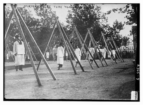 The image is a historical black-and-white photograph depicting a group of individuals in a public setting, possibly during a public execution or a ceremonial event. The text at the top of the image reads "MURDERERS OF TURK WAR MINISTER." The scene is outdoors, with trees and a fence visible in the background, suggesting it is taking place in a public space.
In the foreground, there are multiple wooden structures arranged in a row, which appear to be scaffolds or gallows. Several individuals are standing or walking around these structures. They are dressed in traditional attire, with some wearing long robes and others in simpler clothing. The individuals appear to be observing or participating in the event. The ground is bare, and the overall atmosphere suggests a somber and serious occasion. The photograph has a vintage quality, with a caption and a number (2765-9) in the lower right corner, indicating it is a historical image.