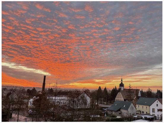 A vibrant sunset fills the sky with shades of orange and pink, featuring textured clouds. In the foreground, buildings and a church with a tower are visible, along with a smokestack and trees.