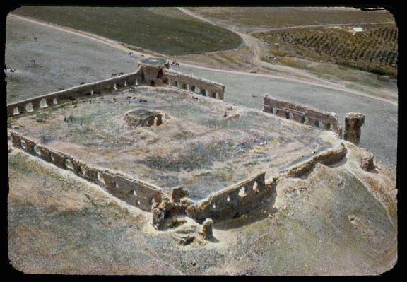 The image shows an aerial view of the ruins of what appears to be a historical structure, possibly from ancient times. The site is rectangular with rounded corners and features several walls that are in various states of preservation. Some sections have standing stone blocks while others show rubble or missing stones.
In the background, there's open land with patches of greenery suggesting fields or cultivated areas. A road runs through the scene, indicating modern accessibility to this archaeological site. The overall color tone is a faded yellowish-brown, indicative of age and weathering on both the ruins and the photograph itself.