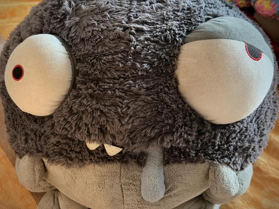 English: A large, grey plush creature with oversized, expressive eyes and a small mouth with teeth. The texture is fuzzy and soft. It is resting on a patterned blanket. 

Deutsch: Eine große, graue Plüschkreatur mit übergroßen, ausdrucksstarken Augen und einem kleinen Mund mit Zähnen. Die Textur ist unscharf und weich. Es ruht auf einer gemusterten Decke.