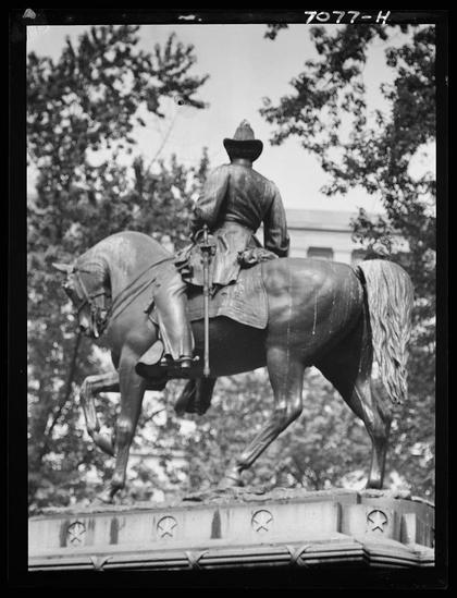 A black and white photograph of a bronze equestrian statue depicting a man in historical attire, mounted on horseback. The figure is likely to be a military or cavalry leader from the American West given his wide-brimmed hat and formal riding gear. This person appears to be galloping forward with one leg raised, suggesting motion within the still sculpture.
The background features trees and what seems to be part of an institutional building, possibly indicating that this statue is located in a park or public square near government structures. The image has visible text "7077-H" at the top corner which may indicate its cataloging number for archival purposes.
