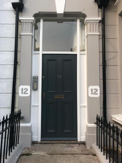 A dark front door with a large glass window above it. A brass number 12 is on the door, and the number 12 has also been stencilled onto the grey pillars surrounding the door.