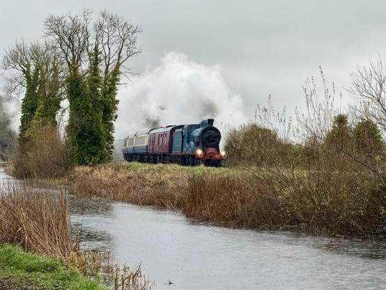 A landscape view of a blue steam train with a big head of clean white steam pulling a burgundy brake van and a rake of blue and cream vintage carriages along the far bank of a rural canal. A few bare trees with plenty of ivy on their trunks separate the railway and the canal. Both are gently curving into view from left to right. The sky overhead is a dull grey and there are rain drops visible on the water.