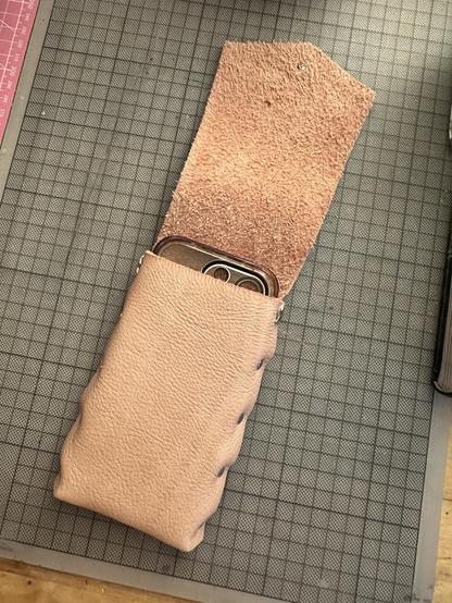Pochette de cuir pile au format de la coque de mon iPhone 14 Pro.
Le cuir est rose. On voit cinq rivets sur chaque côté de la pochette, qui est posée à plat sur mon établi, avec un quadrillage centimétrique.
