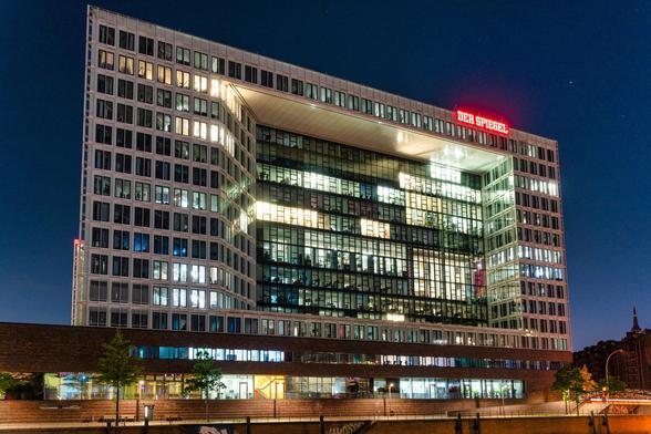 Das Spiegel-Verlagshaus bei Nacht
Das Bild ist eine Nahaufnahme eines großen, modernen Bürogebäudes in der Nacht.
Gebäude: Es handelt sich um das Spiegel-Verlagshaus in Hamburg, erkennbar an der prominenten roten Leuchtschrift "DER SPIEGEL" auf dem Dach.
Architektur: Das Gebäude weist eine auffällige moderne Architektur auf, insbesondere durch die große, viereckige Aussparung in der Mitte der Fassade.
Beleuchtung: Viele Fenster sind beleuchtet, was auf die Büronutzung hindeutet und die Tiefe und Struktur des Inneren durch die Glasfassade sichtbar macht. Der untere Gebäudeteil scheint aus rötlichem Backstein zu bestehen.
Stimmung: Die Beleuchtung der Büros in der klaren, tiefblauen Nacht schafft eine urbane, beschäftigte Atmosphäre.