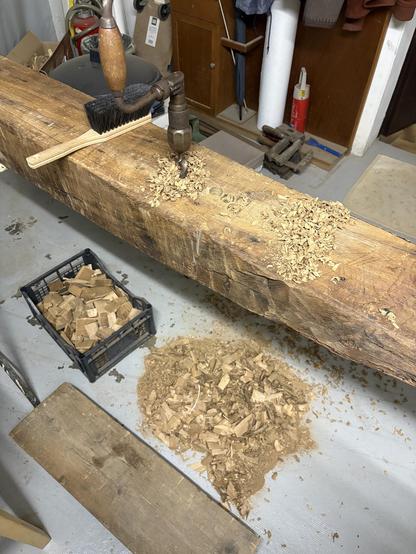 An old barn post with large holes being bored into with a brace and bit. The brace is sticking upright in a hole unattended. Wood chips strewn the post and the floor.