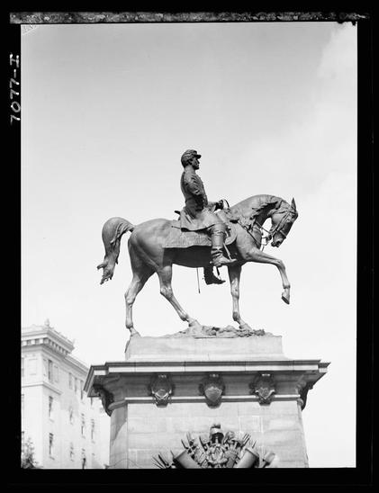 The image depicts a black and white photograph of an equestrian statue on a pedestal, which appears to be made of stone. The statue shows a person dressed in what seems like military attire riding atop a horse that is captured mid-stride with one hoof raised as if galloping or rearing up.
In the background, behind the statue's base, there are parts of classical-style buildings visible, suggesting an urban setting for this monument. Above and around the pedestal, sculptural details such as reliefs can be seen which add to its grandeur but their specific designs aren't clearly discernible in black and white photography.
The sky is clear with no clouds indicating good weather during the time when the photo was taken. The overall composition of the photograph focuses on capturing this significant public sculpture against an urban backdrop, emphasizing both as a tribute or commemoration piece likely related to historical military service given its design elements.