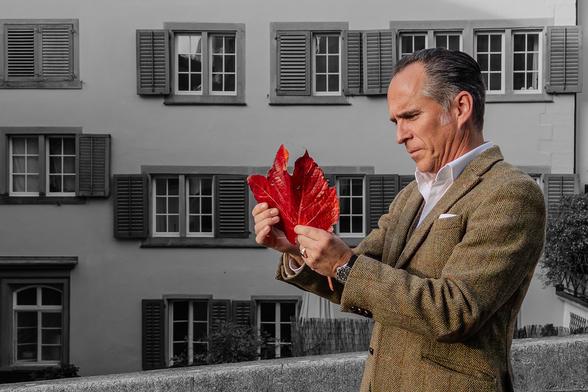 Martin Grassberger steht vor einem Gebäude mit grauen Fensterläden und hält ein leuchtend rotes Rebenblatt in den Händen. Der Hintergrund ist in Schwarz-Weiss gehalten, während Blatt und Kleidung farbig hervorgehoben sind.