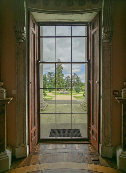 This elegant image captures a view from inside a grand historic building looking out through tall wooden double doors. The doors frame a classical glass-paneled entryway that opens onto a stone terrace, beyond which stretches a manicured formal garden with symmetrical lawns, sculpted topiaries, and mature trees beneath a partly cloudy sky. The interior features ornate architectural details including decorative moldings, curved walls with classical pilasters, and a polished wooden floor that follows the gentle curve of the rounded entrance hall. The composition draws the eye from the weathered grandeur of the interior through multiple frames to the pastoral landscape beyond, creating a sense of depth and the timeless elegance of a stately manor or palace.