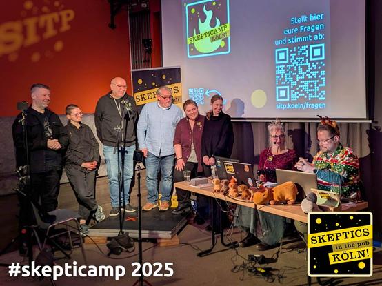 Stehend die sechs Speaker*innen des ersten Deutschen Skepticamps, die fragend die Sabine und Gerald anblicken, die moderiert haben und dabei die ganze Zeit sitzen durften.