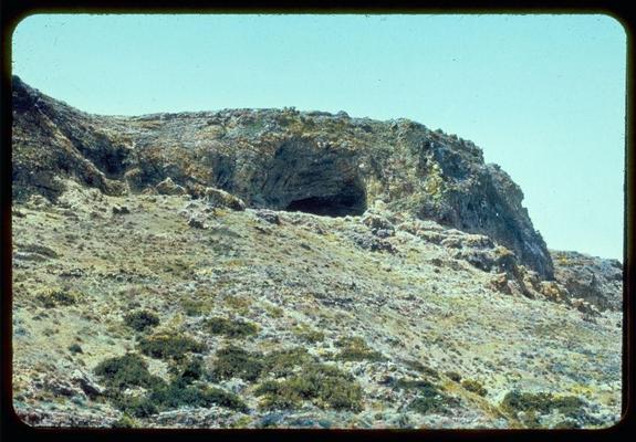 This image depicts a natural landscape characterized by rugged terrain and sparse vegetation. A large, rocky cliff dominates the scene with an opening or cave visible at its top. The surrounding ground is covered in dry grasses, shrubs, and small bushes that indicate arid conditions typical of Mediterranean climates. Above lies a clear blue sky suggesting fair weather during this time frame captured by Matson Photo Service between 1950-77 as per the reference found on Jaffa to Jerusalem.
