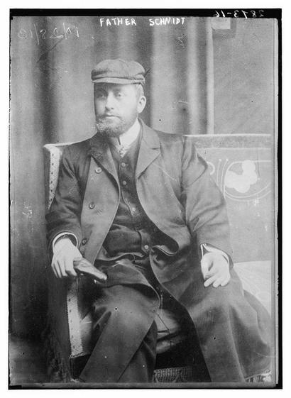 The image is a black-and-white photograph of a man seated on a chair. The man appears to be in formal attire, wearing a long coat or suit with a vest underneath. He has a neatly trimmed beard and mustache, and he is wearing a cap or flat hat. The man is seated with his legs crossed and his hands resting on his lap. The background is simple, with a plain backdrop that appears to be a curtain or a similar material. The photograph has a vintage quality, suggesting it is from an earlier time period. There is text at the top of the image that reads "FATHER SCHMIDT," which may indicate the name of the individual or the subject of the photograph. The image has a rectangular format with a thin black border, and there are some faint markings or text in the top left corner, possibly related to cataloging or identification purposes.