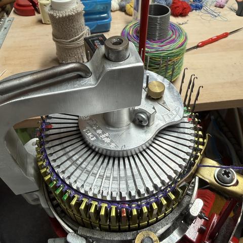 Blick auf eine Sockenstrickmaschine. Viel ist nicht zu sehen, weil die Ribber-Scheibe aufgesetzt ist. Darunter entsteht gerade eine Socke bzw. die Ferse derselben. Blindstricken, yay!