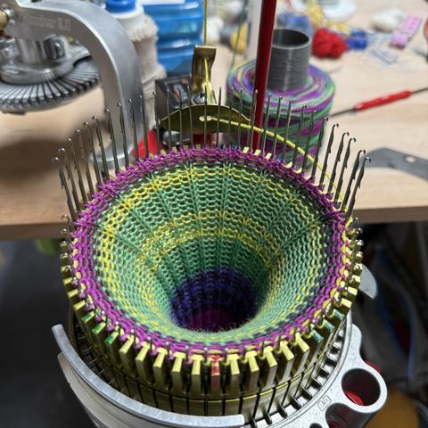 Blick auf die Sockenstrickmaschine, jetzt ohne Ribber. Im Zylinder ist eine fast fertig gestrickte Socke mit Rippenmuster in der hinteren Hälfte.
Das Garn ist grün, gelb, dunkelgrau und pink.