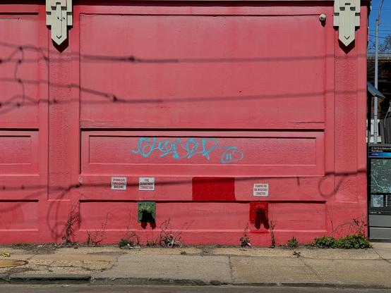A sidewalk runs along the bottom of the photo. Prominently featured is an industrial building’s wall that is painted pink. Shadows of electrical lines snake across the wall. It’s a sunny day.