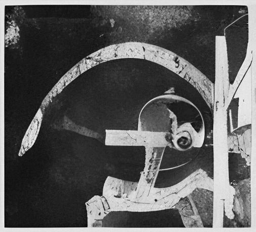 A black and white photograph documenting markings found on a dark asphalt surface. The image shows white paint forming several distinct elements: a large curved arc spanning the upper left portion, straight linear segments creating perpendicular intersections, a circular form containing what appears to be an organic spiral object at its center, and angular directional markings on the right side of the frame. The paint exhibits deterioration with visible cracking, chipping, and areas of loss. The underlying surface shows weathering and textural variations. Dark staining and discoloration are visible across multiple areas of the pavement. The photographic angle is directly overhead, capturing approximately 4-6 feet of surface area.