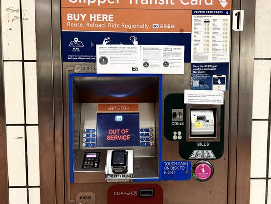 A Clipper Transit Card vending machine is mounted into a tiled subway station wall. The top panel reads “BUY HERE – Reuse. Reload. Ride Regionally.” with icons for different Bay Area transit systems. In the center, a digital screen glows red with the message “OUT OF SERVICE.” Surrounding the screen are metal selection buttons labeled A through H. Below are a keypad and a credit/debit card reader. To the right, a separate payment panel shows labeled slots for COINS and BILLS, with illustrated coin and dollar icons, and a sticker instructing riders to “TOUCH CARD ON DISK TO RIGHT.” Additional notices and fare information posters are taped above and beside the machine, including a Clipper card fares chart. The machine appears worn and scratched, emphasizing its heavy public use.